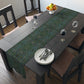 A green floral patterned table runner inspired by William Morris, laid out on a wooden dining table with plates and cutlery set on top.