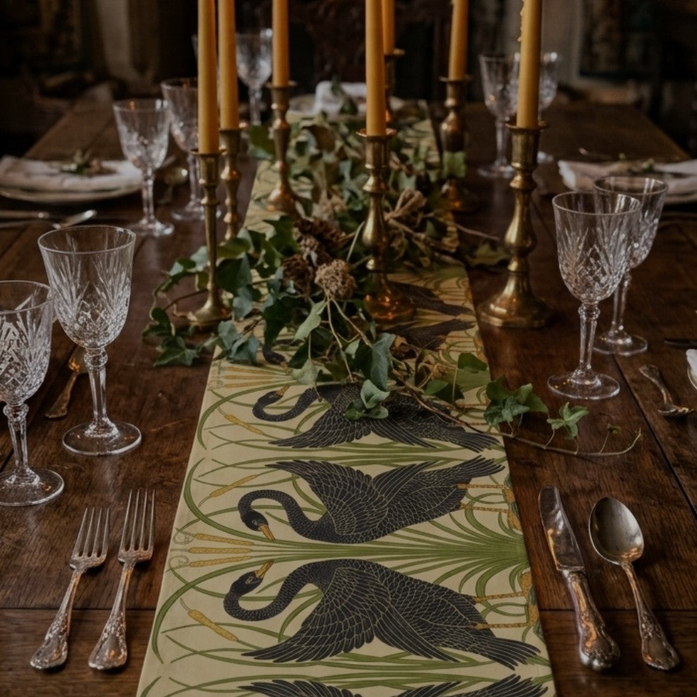 A decorative table runner with a black crane pattern on a light beige background, styled on a dark wooden dining table with crystal glasses, silverware, candles, and greenery.