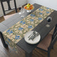A table runner with a Victorian floral design in blue and green on a beige background, laid out on a dining table with place settings and decorative flowers.