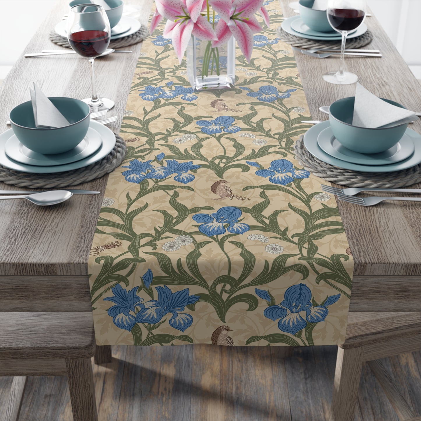 A table runner with a Victorian floral design in blue and green on a beige background, laid out on a dining table with place settings and decorative flowers.