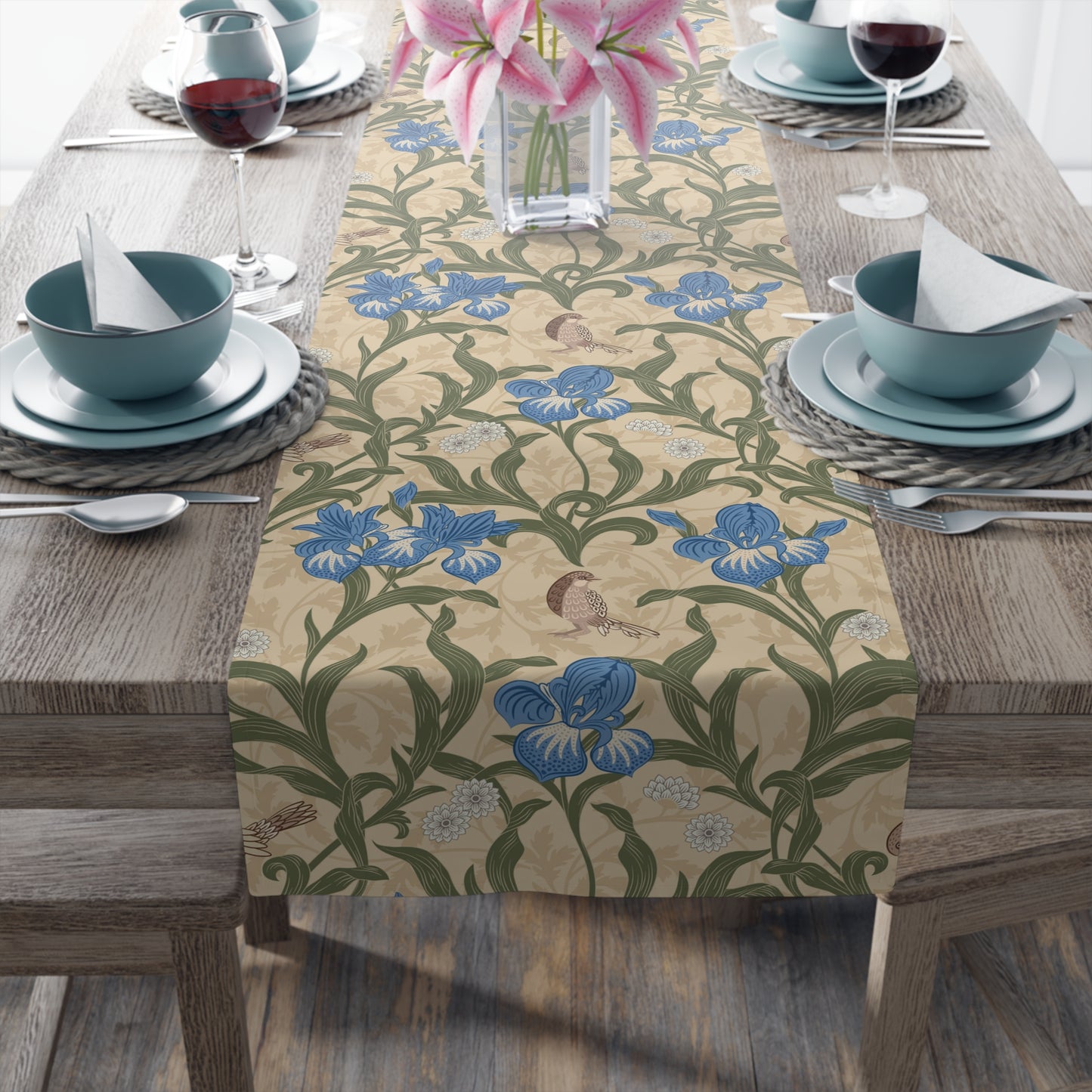 A table runner with a Victorian floral design in blue and green on a beige background, laid out on a dining table with place settings and decorative flowers.