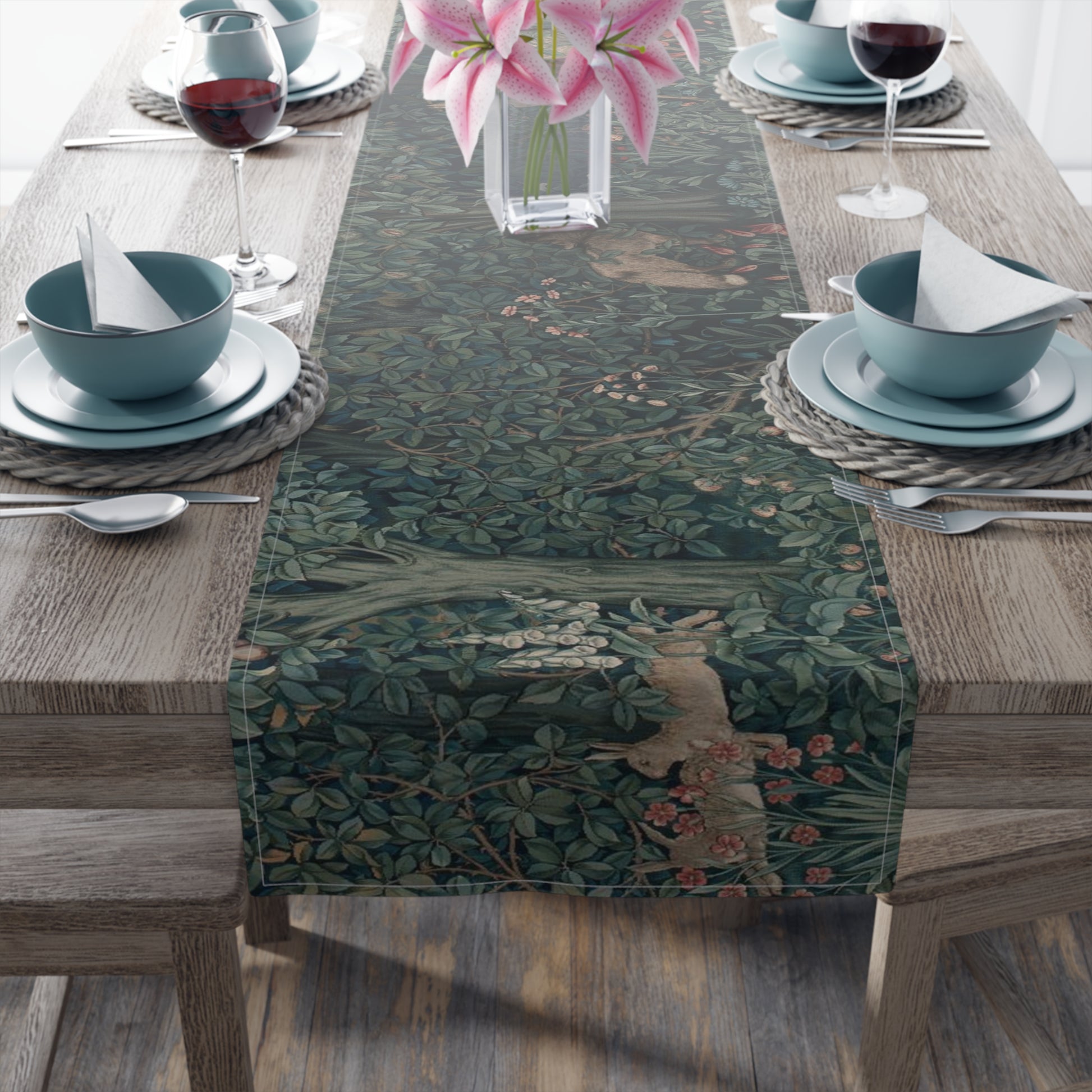 A table runner with a floral William Morris & Co pattern, featuring green leaves and pink flowers, laid out on a wooden dining table with cutlery and a vase of pink flowers in the background.