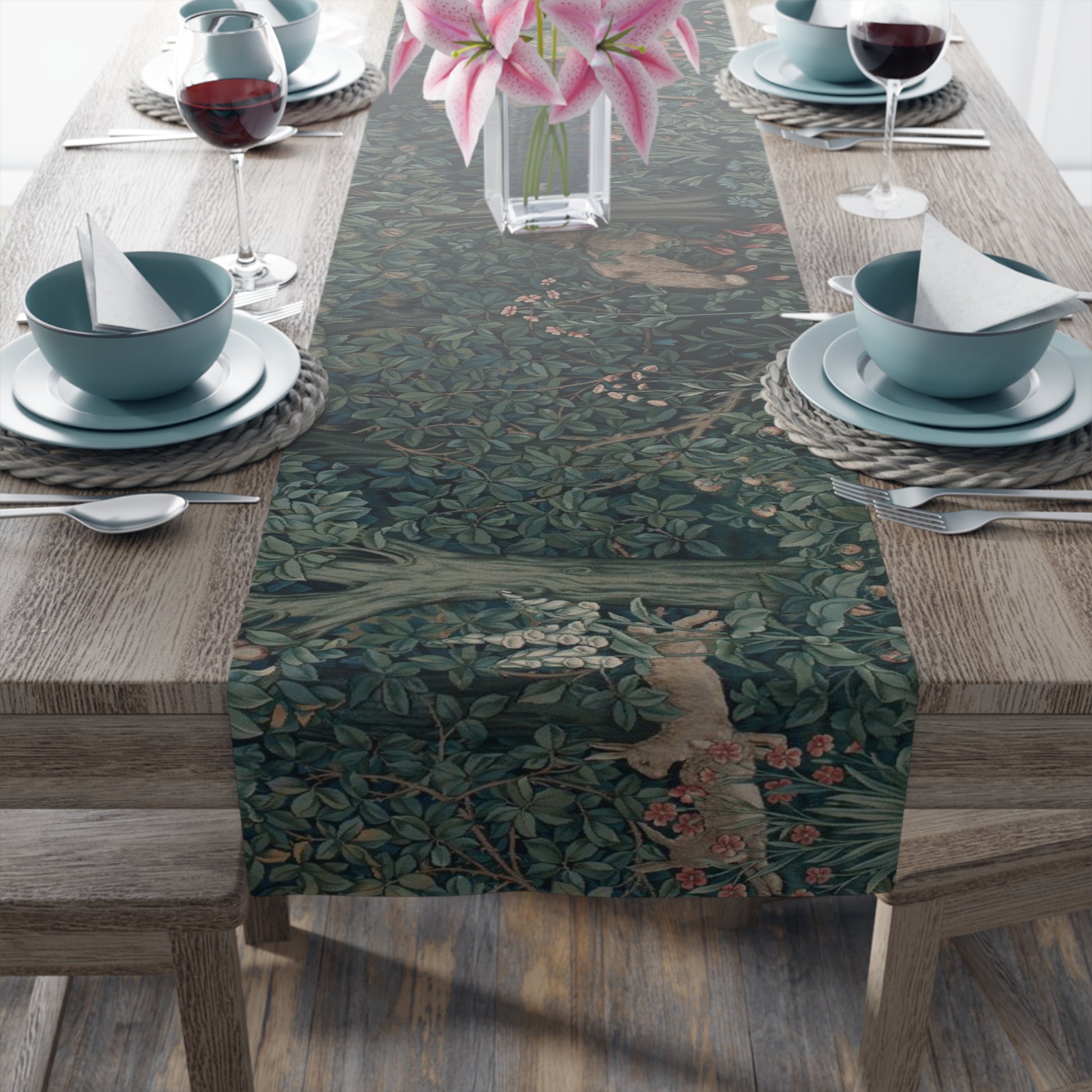 A table runner with a floral pattern, featuring green leaves and pink flowers, laid out on a wooden dining table with cutlery and a vase of pink flowers in the background.