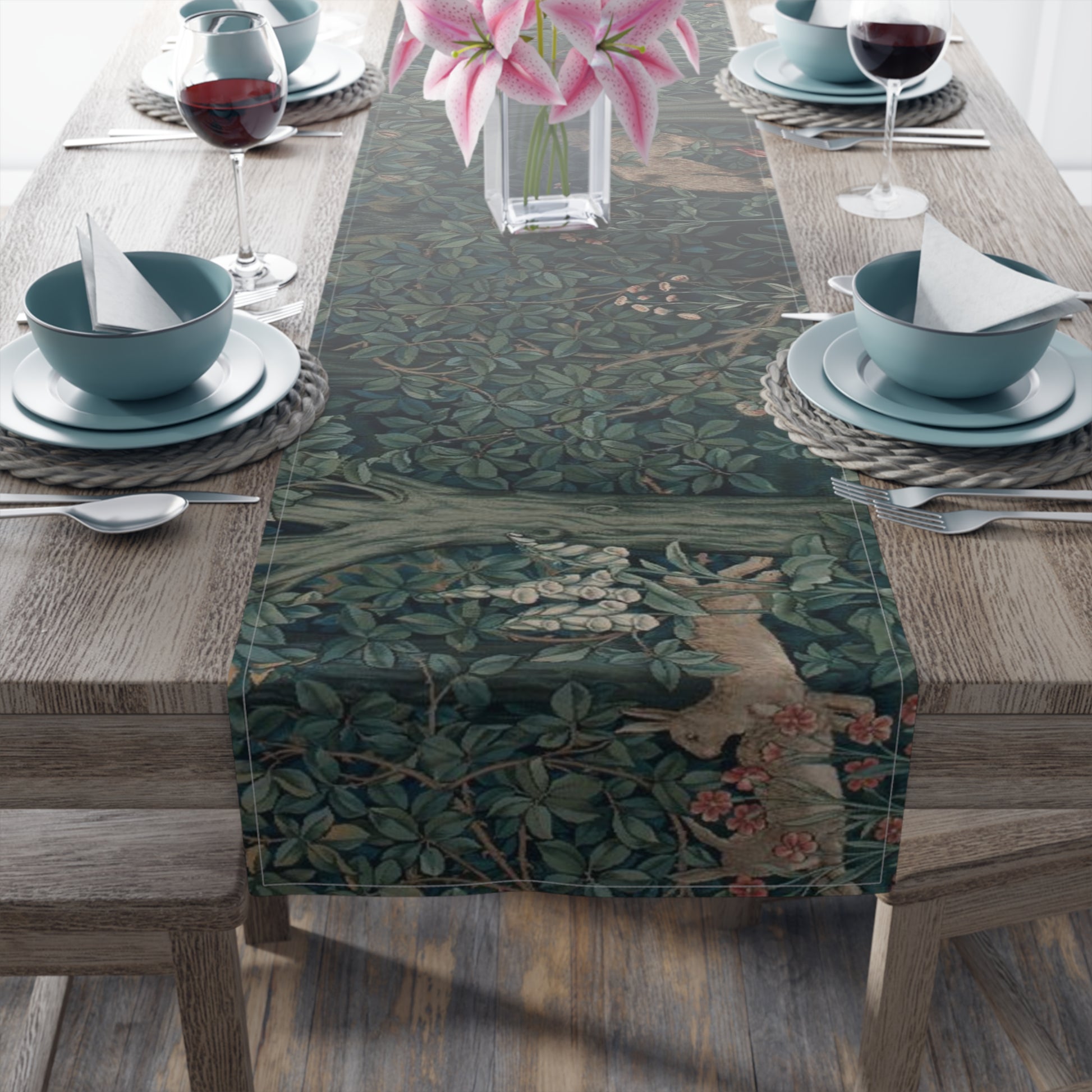 A table runner with a floral pattern, featuring green leaves and pink flowers, laid out on a wooden dining table with cutlery and a vase of pink flowers in the background.