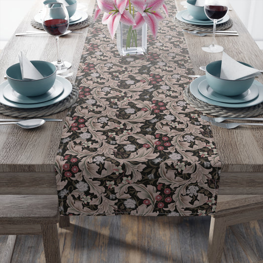 A table runner with a William Morris & Co design laid out on a dining table, flanked by plates and a vase with flowers, set for a meal.