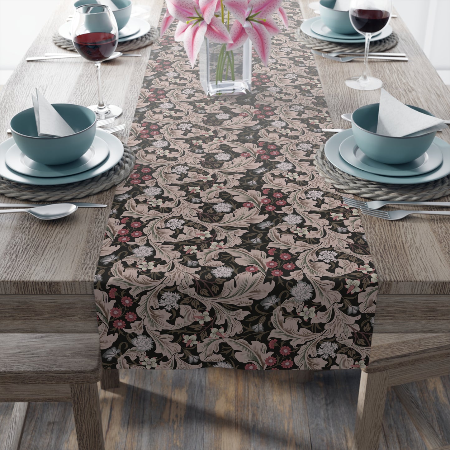 A table runner with a William Morris & Co design laid out on a dining table, flanked by plates and a vase with flowers, set for a meal.