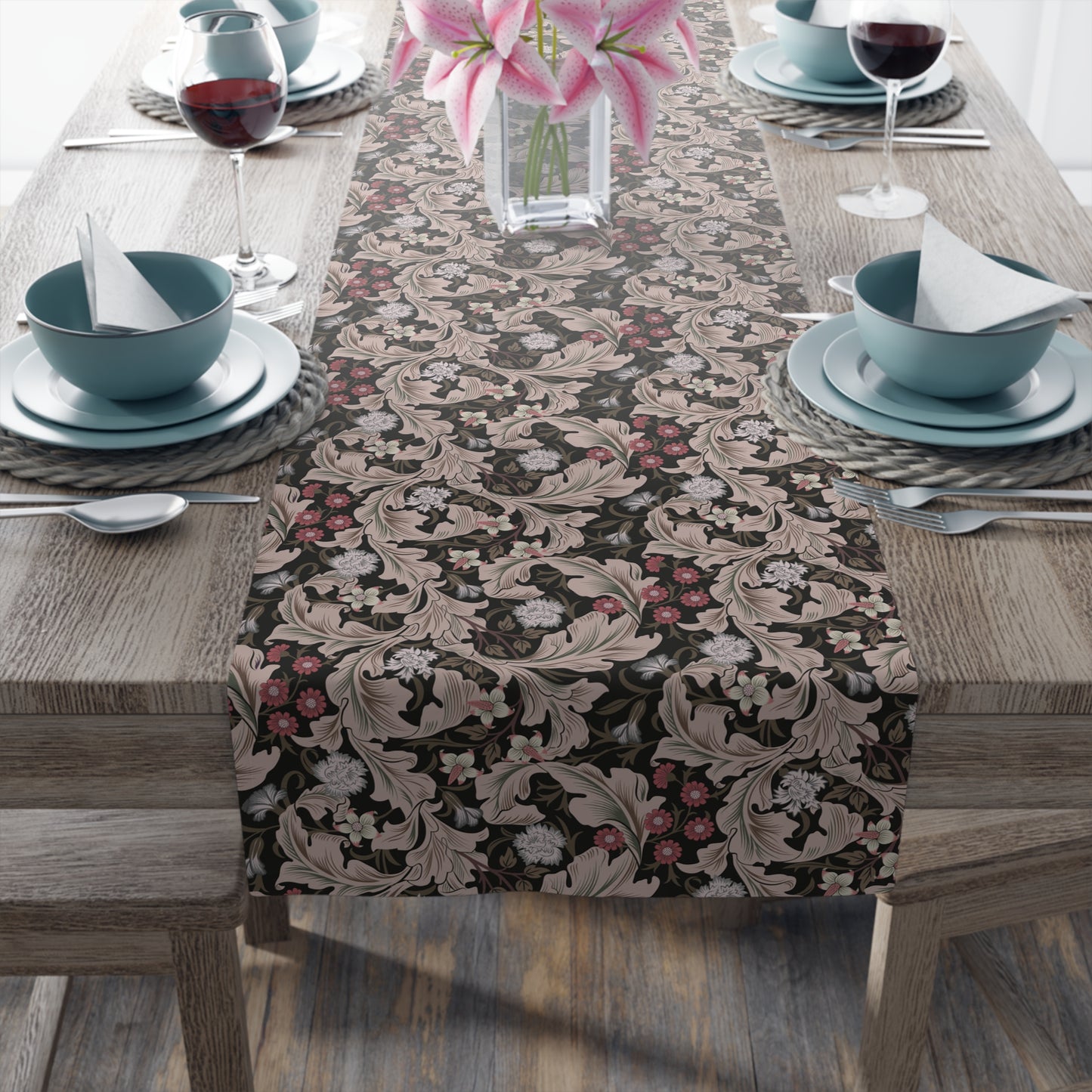 A table runner with a William Morris & Co design laid out on a dining table, flanked by plates and a vase with flowers, set for a meal.