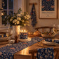 A styled dining table at night with a navy floral table runner, matching napkins, a lit floral candle, a white pitcher of flowers, bread, plates, and wine glasses. A framed William Morris & Co print hangs on the wall behind.