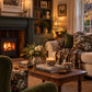 A cozy, warmly lit living room featuring a dark green fireplace mantel with a burning wood stove, a cream sofa adorned with floral throw pillows and a matching patterned blanket, a wooden coffee table with books and candles, and a window with sheer curtains.