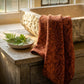 A terracotta-colored bath towel with an embossed botanical leaf pattern draped over a rustic wooden table next to a stone tub and a small bowl of green ferns.