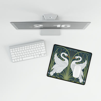 A rectangular desk mat with a floral and swan William Morris & Co pattern design, featuring a white background and swans, placed under a white mouse pad with a keyboard on top.