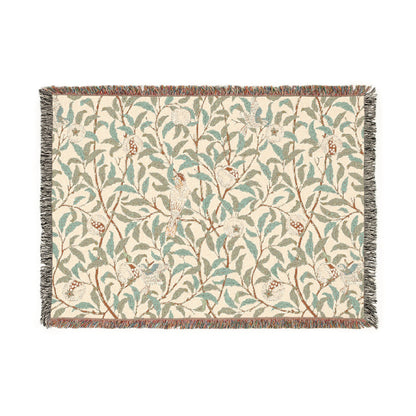 A woven cotton blanket with a geometric bird and pomegranate William Morris & Co pattern in beige, green, and brown colours, with fringed edges.