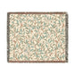 A woven cotton blanket with a geometric bird and pomegranate pattern in beige, green, and brown colors, with fringed edges.