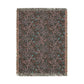 A rectangular woven throw blanket with fringed edges, featuring an abstract floral pattern in dusty pink, teal blue, and light gray on a dark charcoal background.
