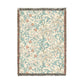 A rectangular woven throw blanket with a cream background, featuring a detailed botanical and animal pattern in shades of teal, dusty pink, and beige, finished with dark gray and brown fringe on all sides.