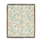 A rectangular woven throw blanket with a cream background and an intricate botanical pattern in teal, dusty pink, and beige, finished with dark gray and rust fringe on all sides.