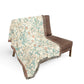 A woven throw blanket with a detailed botanical pattern draped over a brown armchair, shown against a plain white background.