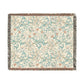 A square woven throw blanket with a cream background and an intricate floral and vine pattern in light blue, beige, and muted coral. The blanket is finished with dark gray and brown fringed edges.