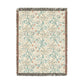 A rectangular woven throw blanket with a cream background and an all-over botanical pattern in muted teal, dusty orange, and beige, finished with dark gray and brown fringe on all sides.