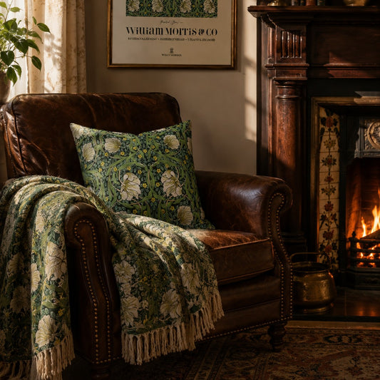 A cozy living room scene featuring a brown leather armchair with a green and cream floral patterned throw blanket draped over it and a matching decorative pillow. A lit fireplace and framed art are visible in the background.