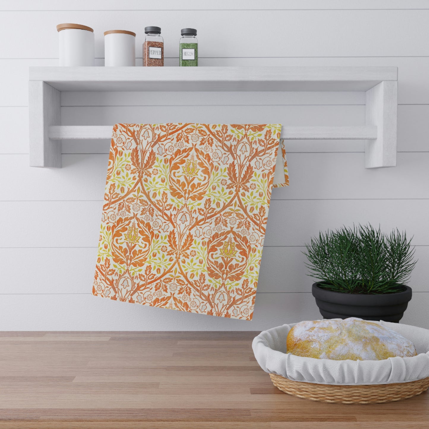 A kitchen tea towel with a floral William Morris  & Co pattern in orange, green, and yellow colours on a light background. 