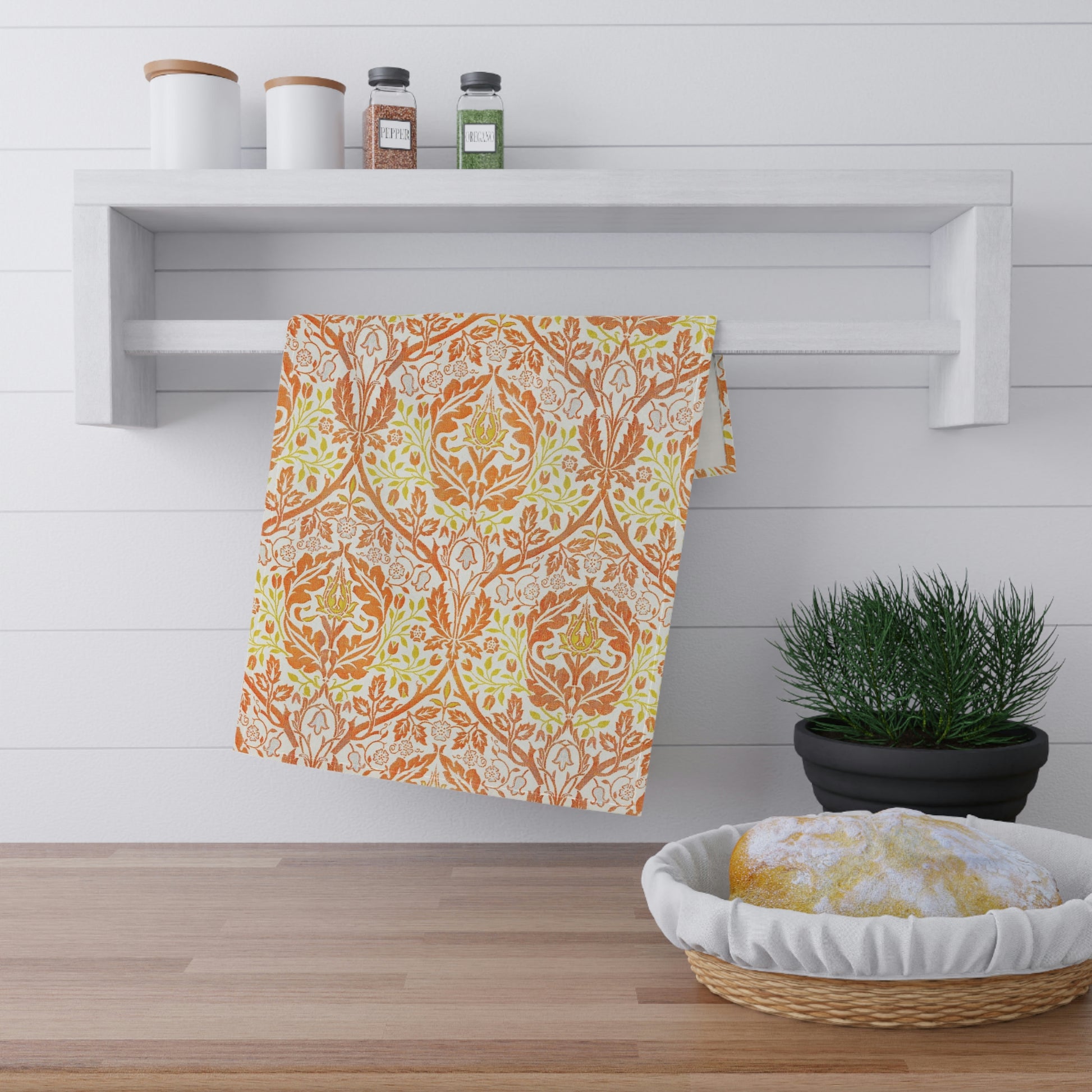 A kitchen tea towel with a floral William Morris  & Co pattern in orange, green, and yellow colours on a light background. 