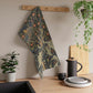 A kitchen tea towel featuring an artistic William Morris & Co design with birds, flowers, and leaves on a green background.